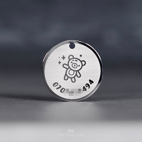 Rund silvrig ID-bricka, namnbricka med personlig gravyr, telefonnummer och ikon på en nalle