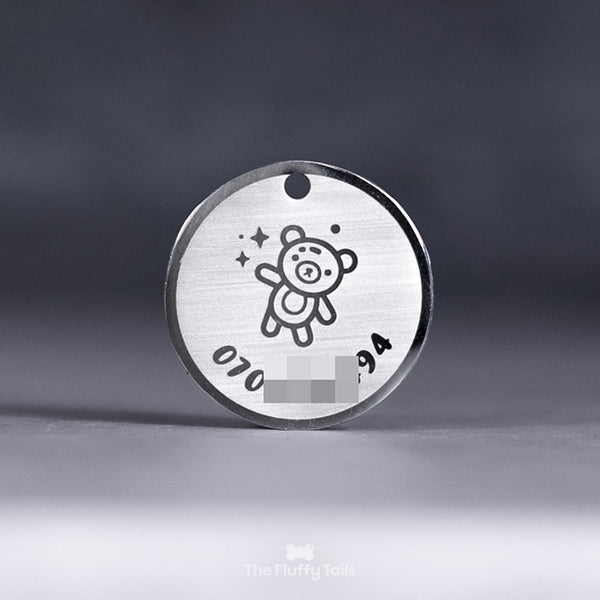 Rund silvrig ID-bricka, namnbricka med personlig gravyr, telefonnummer och ikon på en nalle