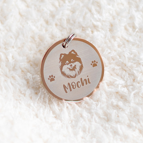 Rund ID-bricka i roséguld på vitt fluffigt underlag, med gravyr på hundrasen pomeranian, två tassar på varje sida och hundens namn