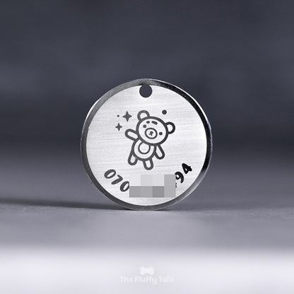 Rund silvrig ID-bricka, namnbricka med personlig gravyr, telefonnummer och ikon på en nalle