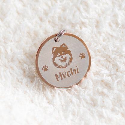 Rund ID-bricka i roséguld på vitt fluffigt underlag, med gravyr på hundrasen pomeranian, två tassar på varje sida och hundens namn 