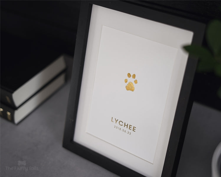 Tassavtryck i guldigt folierat tryck, inramad i en svart ram med passpartout, ståendes på en grå yta