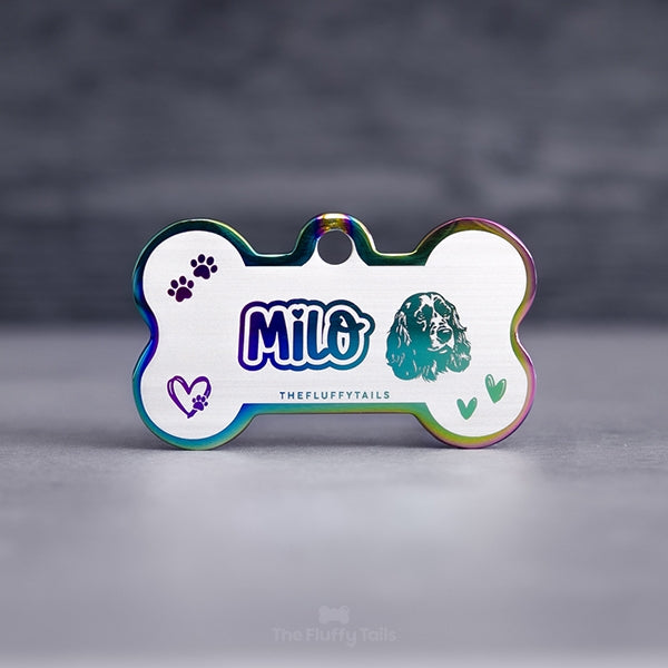 Hundbensformad flerfärgad namnbricka i regnbågens färger, graverad med namn i lekfullt typsnitt med ett hjärta över i:et, ikoner på två handritade hjärtan, tassavtryck och en hund av hundrasen cocker spaniel. Namnbrickor passar även katter.