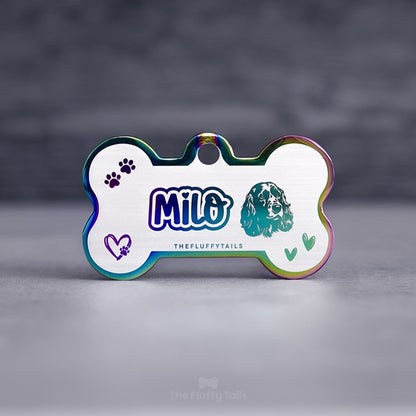 Hundbensformad flerfärgad namnbricka i regnbågens färger, graverad med namn i lekfullt typsnitt med ett hjärta över i:et, ikoner på två handritade hjärtan, tassavtryck och en hund av hundrasen cocker spaniel. Namnbrickor passar även katter.