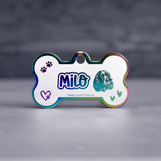 Hundbensformad flerfärgad namnbricka i regnbågens färger, graverad med namn i lekfullt typsnitt med ett hjärta över i:et, ikoner på två handritade hjärtan, tassavtryck och en hund av hundrasen cocker spaniel. Namnbrickor passar även katter.