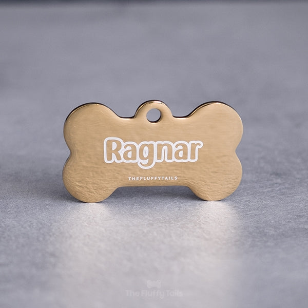 Hundbensformad guldfärgad namnbricka, graverad med namn i enkel kontur och handskrivet typsnitt