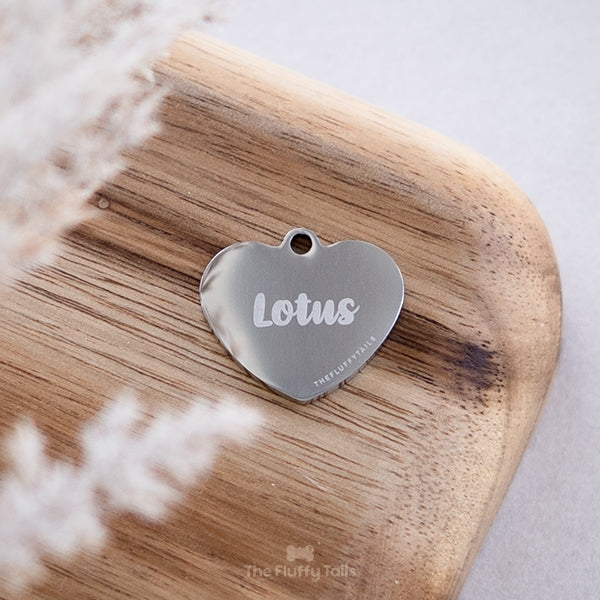 Silvrig hjärtformad namnbricka graverad med namn i skrivstil, placerad på en bricka i akaciaträ