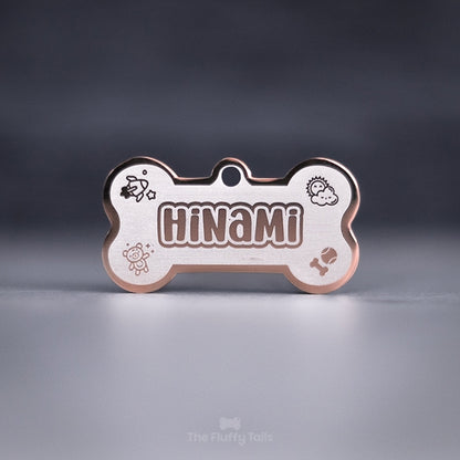 Hundbensformad namnbricka i färgen roséguld, tillverkad av rostfritt stål, graverad med personlig design som består av hundens namn, detaljer och ikoner på en raket, sol, nallebjörn, boll och tuggben