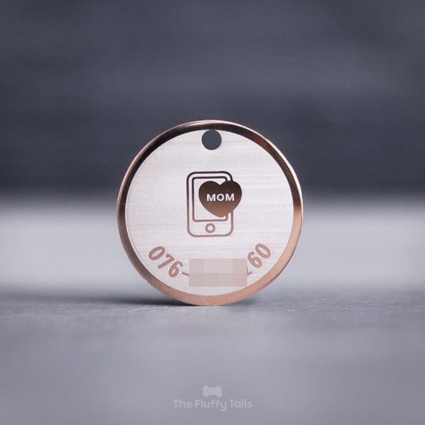 Rund namnbricka i färgen roséguld, för både hund och katt. Graverad med hundens ägares telefonnummer och en ikon i mitten på en smartphone och ett hjärta med texten mom. 