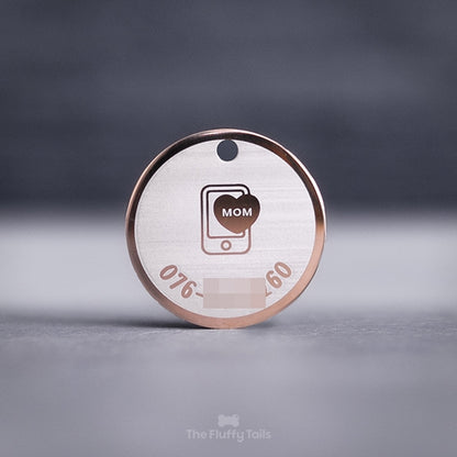 Rund namnbricka i färgen roséguld, för både hund och katt. Graverad med hundens ägares telefonnummer och en ikon i mitten på en smartphone och ett hjärta med texten mom. 