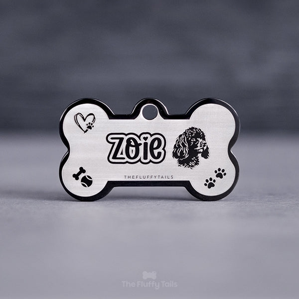 Svart hundbensformad namnbricka graverad med graverad med namn i ett lekfullt typsnitt med hjärta över i:et och konturer runt texten, ikoner av hjärta med tass, tennisboll, tuggben och tassavtryck.