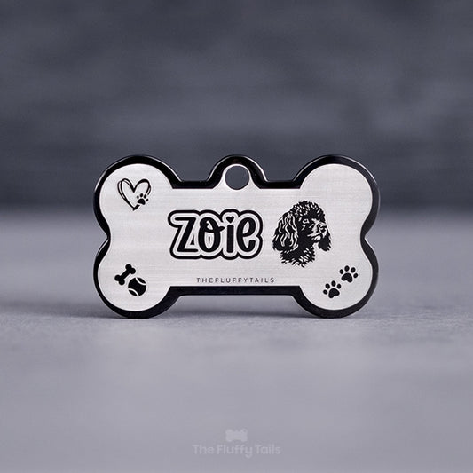 Svart hundbensformad namnbricka graverad med graverad med namn i ett lekfullt typsnitt med hjärta över i:et och konturer runt texten, ikoner av hjärta med tass, tennisboll, tuggben och tassavtryck.
