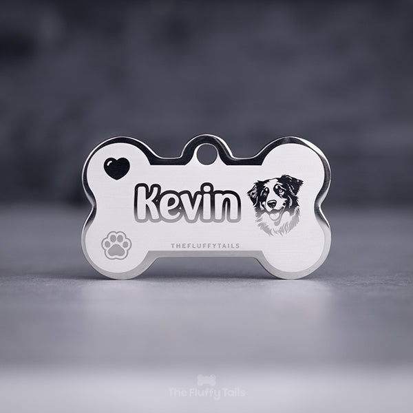 Silvrig hundbensformad namnbricka av rostfritt stål, graverad med hundens namn i enkel kontur och ikoner på ett hjärta, en tass och en hund av hundrasen australian shepherd.