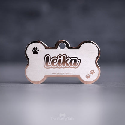 Roséfärgad, hundbensformad namnbricka av rostfritt stål för hundar och katter, graverad med djurets namn i skrivstil med dubbelkontur och tassavtryck.
