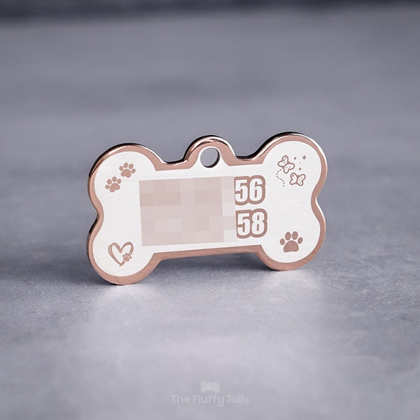 Baksidan på en roséfärgad, hundbensformad namnbricka av rostfritt stål för hundar och katter, graverad med två telefonnummer till ägarna med kontur och fyra ikoner av tassar, fjärilar med glitter och hjärta med tassavtryck inuti.