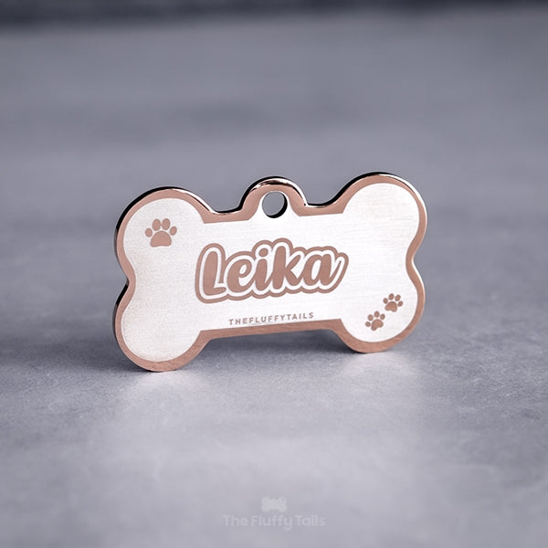 Roséfärgad, hundbensformad namnbricka av rostfritt stål för hundar och katter, graverad med djurets namn i skrivstil med dubbelkontur och tassavtryck