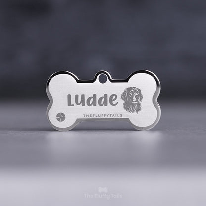 Silvrig hundbensformad, egendesignad namnbricka av rostfritt stål, graverad med hundens namn och en tennisboll samt en hund av hundrasen röd golden retriever.