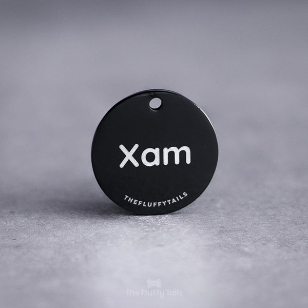 Svart, rund namnbricka av rostfritt stål, graverad med namnet Xam