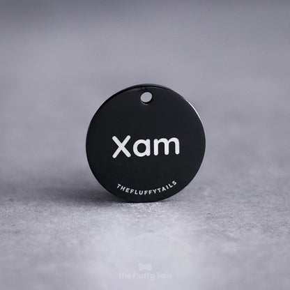 Svart, rund namnbricka av rostfritt stål, graverad med namnet Xam