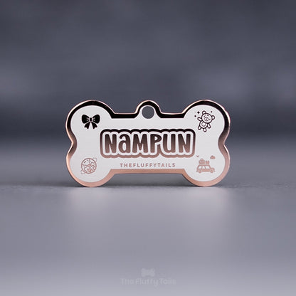 Egendesignad hundbensformad namnbricka, för hund och katt i rostfritt stål i färgen roséguld med egen design, graverad med hundens namn i lekfullt typsnitt samt ikoner av rosett, nalle, jordglob och campingvan