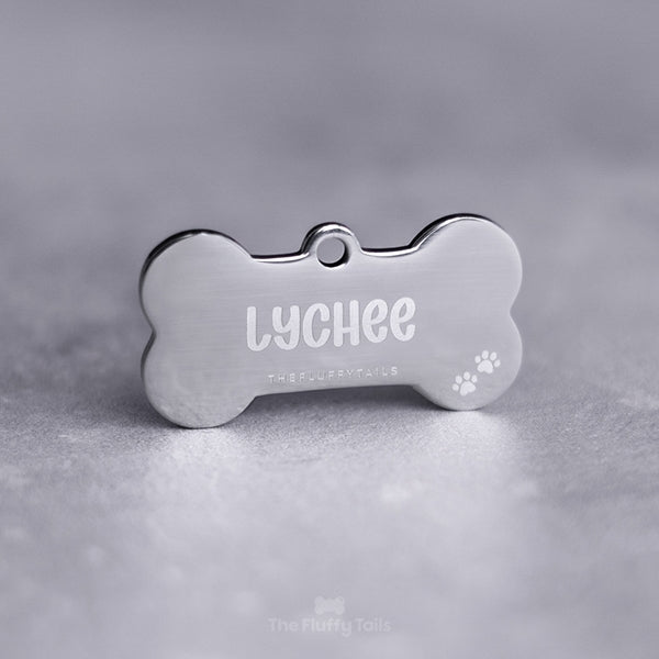 Silvrig, hundbensformad namnbricka av rostfritt stål, graverad med hundens namn och två tassavtryck i högra nedre hörnet