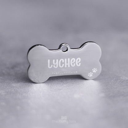 Silvrig, hundbensformad namnbricka av rostfritt stål, graverad med hundens namn och två tassavtryck i högra nedre hörnet