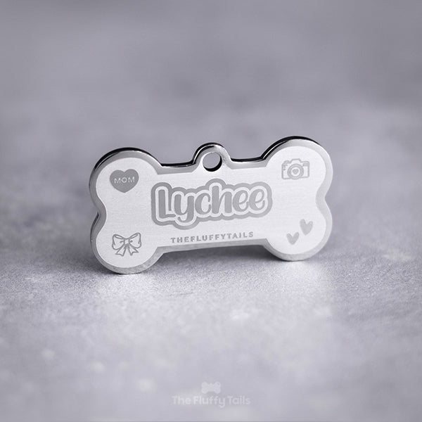 Silvrig hundbensformad namntag, id-bricka, namnbricka av rostfritt stål med egen design och personlig gravyr. Namnbrickan är lasergraverad med hundens namn i dubbelkontur och lekfullt typsnitt, ikoner på ett hjärta med texten mom, kamera, rosett och två handritade hjärtan. Passar även katter. 