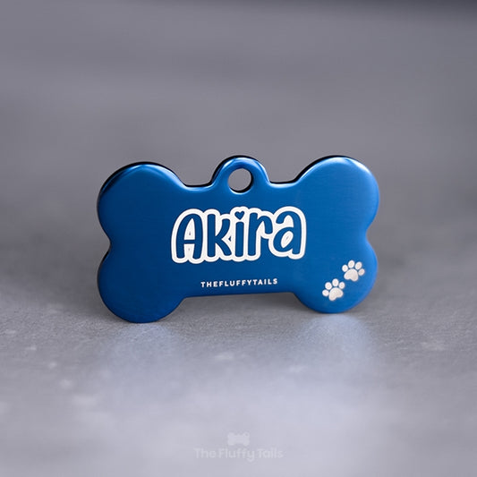 En blå hundsformad namnbricka av rostfritt stål med exklusiv spegelpolering. ID-brickan är graverad med hundens namn i ett lekfullt typsnitt med enkel kontur samt två tassar i högra nedre hörnet.