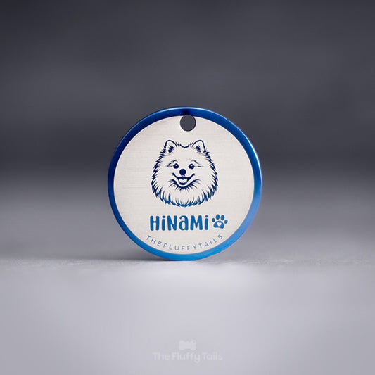 Blå, rund namnbricka av rostfritt stål med personlig gravyr och egen design. Namnbrickan är graverad med en pomeranian hund, hundens namn samt ett bredvid namnet tassavtryck med ett hjärta inuti