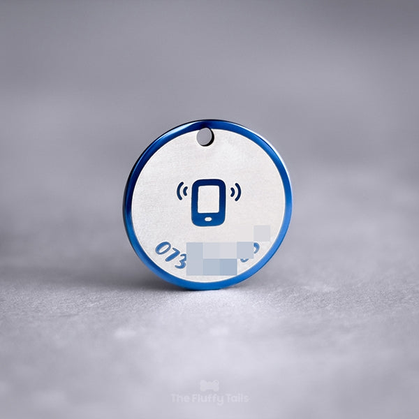 En blå, rund ID-bricka av rostfritt stål med spegelpolering som skiftar fint i ljuset. ID-brickan är graverad med ett telefonnummer längs namnskyltens runda form. I mitten av namnbrickan har en ikon på en ringande telefon graverats. Passar både hundar och katter.