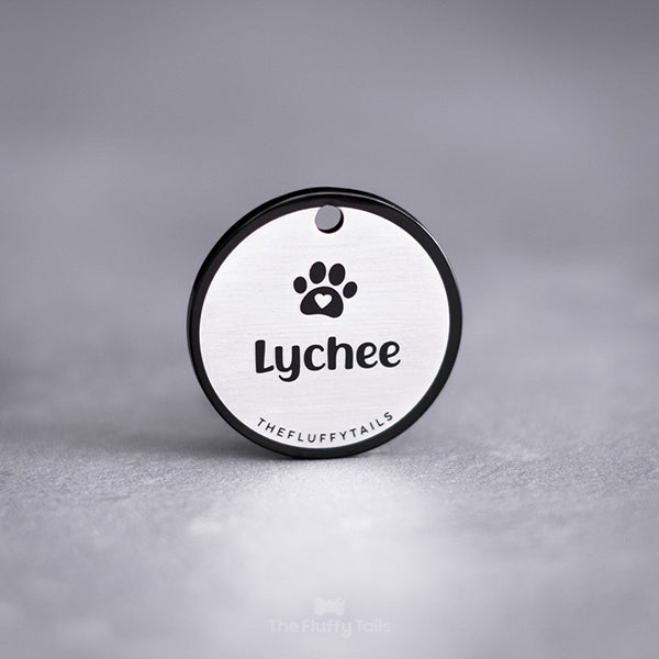 Svart, rund namnbricka av rostfritt stål graverad med egen design förställande hundens namn och en tass med ett hjärta. Namnbrickan passar både hund och katt.