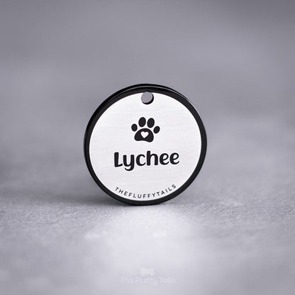 Svart, rund namnbricka av rostfritt stål graverad med egen design förställande hundens namn och en tass med ett hjärta. Namnbrickan passar både hund och katt.