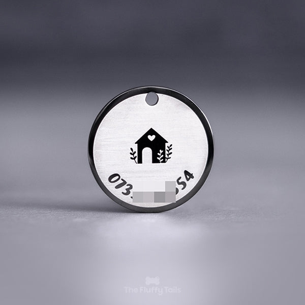 Baksidan på en exklusiv svart, rund namnbricka av rostfritt stål graverad med ett hus och ett hjärta med lite växter. Motivet är placerat i mitten på brickan. Ett telefonnummer till ägaren är graverad längs brickans runda form.