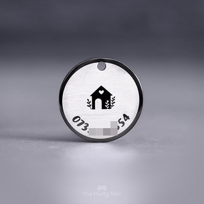 Baksidan på en exklusiv svart, rund namnbricka av rostfritt stål graverad med ett hus och ett hjärta med lite växter. Motivet är placerat i mitten på brickan. Ett telefonnummer till ägaren är graverad längs brickans runda form.