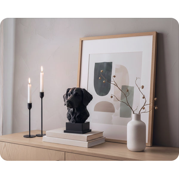 Interiörbild för inspiration med en svart hundskulptur i 3D av hundrasen stabijhoun som står på två böcker, inredd i minimalistiskt och skandinaviskt stil tillsammans med två långa, svarta ljusstakar, tavla i ek, vit hög vas med torkade växter 