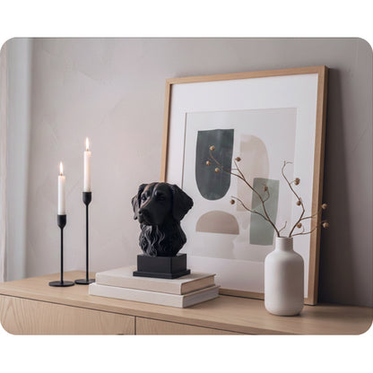 Interiörbild för inspiration med en svart hundskulptur i 3D av hundrasen stabijhoun som står på två böcker, inredd i minimalistiskt och skandinaviskt stil tillsammans med två långa, svarta ljusstakar, tavla i ek, vit hög vas med torkade växter 